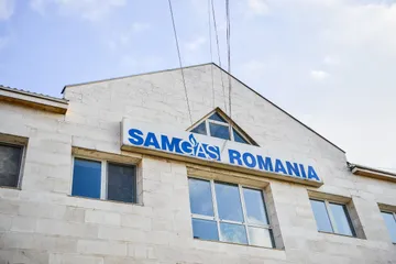 Fațada unei clădiri din piatră de culoare deschisă, cu inscripția albastră SAMGAS ROMANIA deasupra ferestrelor de la etajul superior, pe fundalul unui cer parțial înnorat.