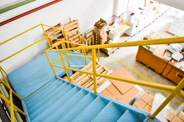 O scară metalică albastră cu balustrade galbene, văzută de sus, care coboară spre pardoseala unui depozit cu paleți de lemn și cutii de carton.