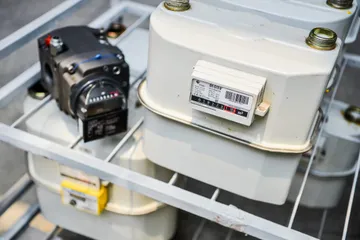 Un contor de gaz alb, cu etichetă și mecanism indicator cu cifre, lângă un contor de gaz negru cu pistoane rotative și mecanism indicator mecanic, ambele pe un stelaj metalic alb.