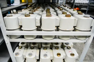 Rânduri de corpuri albe de contor de gaz cu racorduri filetate de alamă, pe rafturile unui stelaj metalic gri.