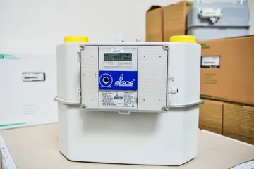 Un contor de gaz inteligent alb, cu afișaj și un panou albastru ELGAS pe partea frontală și capace galbene pe cele două racorduri, așezat în fața unor cutii de carton.