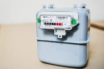 Un contor de gaz gri cu etichetă Pietro Fiorentini și un șir de cifre între două urechi verzi pe partea frontală, așezat pe carton.