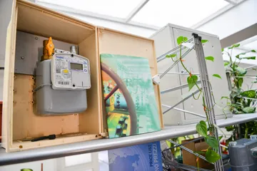 Un contor de gaz inteligent cu afișaj și un robinet de alamă cu mâner portocaliu, într-o carcasă deschisă, de culoare bej, lângă o carte așezată vertical pe un raft, printre plante verzi.