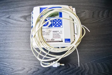 Un convertor ELCOR de volum pentru gaz, cu taste albastre și o etichetă ELGAS, cu cablurile gri strânse în colac deasupra lui, pe o suprafață de lemn închis la culoare.