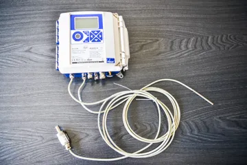 Un convertor electronic ELCOR de volum pentru gaz, cu tastatură albastră și afișaj, cu cabluri gri care se termină cu un senzor de presiune și o sondă de temperatură, pe o suprafață de lemn închis la culoare.