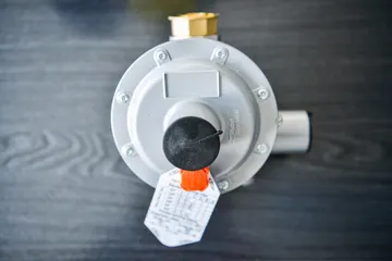Un regulator gri de presiune pentru gaz, văzut dinspre capacul rotund al membranei, cu un căpăcel negru și o etichetă de hârtie, pe o suprafață de lemn închis la culoare.