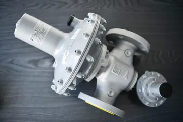 Un regulator gri de presiune pentru gaz, cu o carcasă rotundă a membranei prinsă cu șuruburi și racorduri cu flanșă, așezat pe o suprafață de lemn închis la culoare, lângă un dispozitiv mai mic cu capac negru.