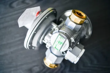 Un regulator gri de presiune pentru gaz, cu racorduri filetate de alamă și o etichetă verde, văzut de sus, pe o suprafață de lemn închis la culoare.