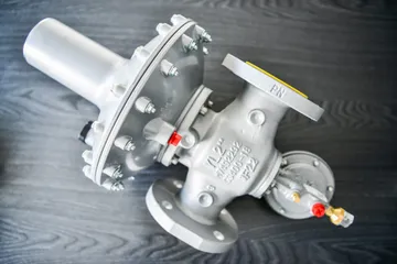 Un regulator gri de presiune pentru gaz, cu flanșe, cu o carcasă mare a membranei prinsă cu șuruburi și un pilot mic cu capac roșu, pe o suprafață de lemn închis la culoare.