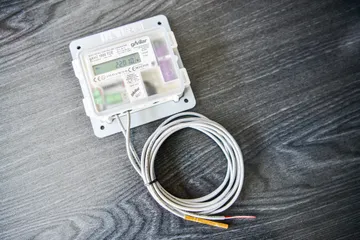 Un convertor electronic de volum pentru gaz, cu un afișaj mic, într-o carcasă albă cu capac transparent, cu un cablu gri strâns în colac și o sondă de temperatură, pe o suprafață de lemn închis la culoare.
