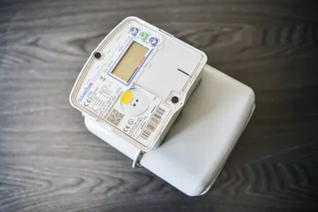 Un contor de gaz inteligent alb, cu afișaj LCD și un buton galben pe panoul frontal, pe o suprafață de lemn închis la culoare.