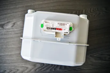 Un contor de gaz cu membrană, alb, cu etichetă Pietro Fiorentini și mecanism indicator mecanic, așezat pe o suprafață de lemn închis la culoare.
