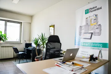 Un birou cu o masă de lucru din lemn deschis la culoare, pe care se află un laptop și hârtii, un scaun de birou negru și, lângă masă, un banner Pietro Fiorentini „Smart Metering”.