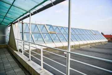 O pasarelă cu balustrade albe, acoperită cu o plasă verde de umbrire, lângă un luminator lung de sticlă pe un acoperiș plat.