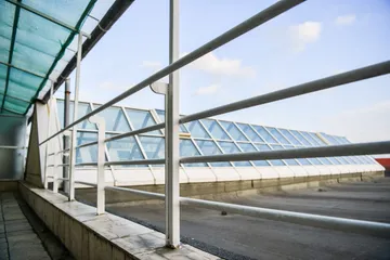 Un luminator lung de acoperiș, văzut printre balustradele albe ale unei pasarele de pe acoperiș, cu o copertină din plasă verde de umbrire.