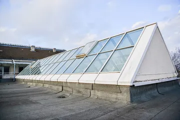 Un luminator lung de sticlă în două ape, cu un fronton alb la capăt, pe un acoperiș plat, pe fundalul unui cer albastru pal.