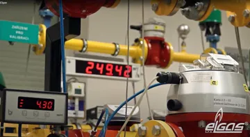 O instalație de etalonare a contoarelor de gaz, cu conducte galbene și roșii, două afișaje digitale roșii și un contor cu partea superioară argintie, legat printr-un cablu albastru, cu sigla ELGAS în colțul din dreapta jos.