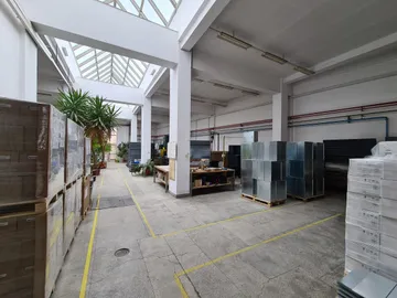 O hală de producție sub un acoperiș de sticlă, cu paleți cu cutii înfoliate în stânga, carcase din tablă stivuite în centru și cutii albe în dreapta.