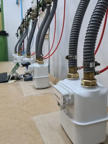Un șir de contoare de gaz albe pe un banc acoperit cu carton, fiecare legat la conductele de pe perete prin furtunuri gofrate gri, cu o tastatură și o tabletă în stânga.