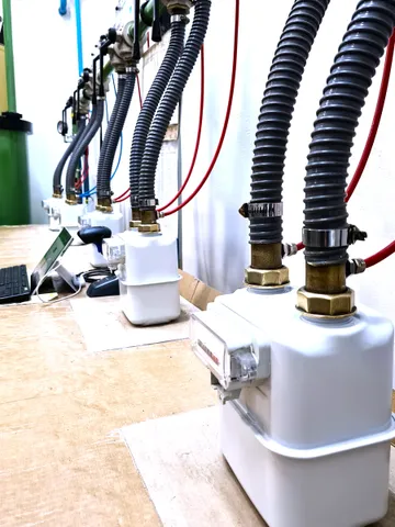 Un șir de contoare de gaz albe pe un banc acoperit cu carton, fiecare legat la conductele de pe perete prin furtunuri gofrate gri, cu o tastatură și o tabletă în stânga.