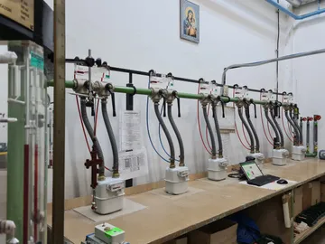 Un stand de verificare de-a lungul unui perete alb, cu un șir de contoare de gaz, fiecare legat prin furtunuri gofrate la o conductă verde, cu un laptop pe stand și o icoană înrămată pe perete, deasupra.