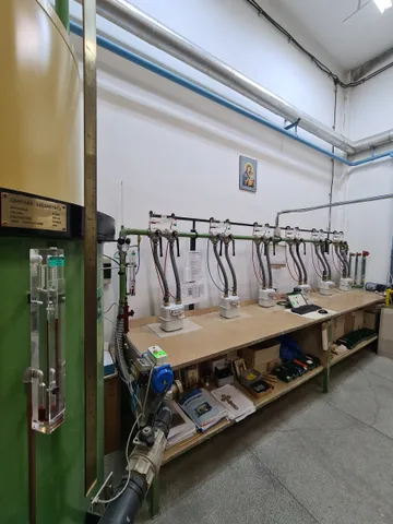 Un stand de verificare a contoarelor de gaz, văzut pe lângă o coloană verde cu un indicator cu tub de sticlă, cu contoare pe stand legate prin furtunuri la conducta de pe perete și cu scule și bibliorafturi pe raftul de dedesubt.