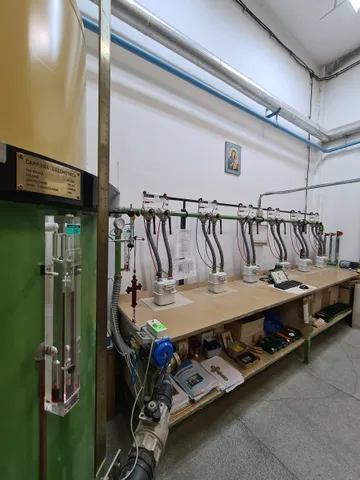 Un stand de verificare a contoarelor de gaz, cu contoare legate prin furtunuri la o conductă de-a lungul peretelui, văzut pe lângă o coloană verde cu o plăcuță de alamă, în prim-plan.
