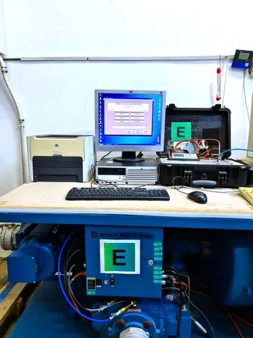 O masă de lucru cu un monitor de calculator, o imprimantă, o tastatură și o valiză neagră deschisă cu un aparat portabil, deasupra unui echipament albastru cu eticheta Model 5 ROOTS Prover.