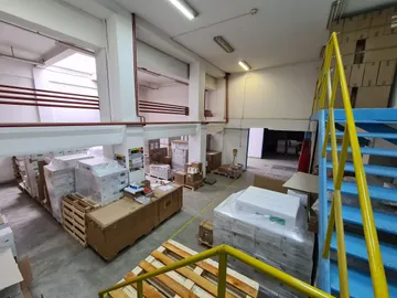 Un depozit văzut de la balustrada galbenă a unei scări, lângă o scară albastră, cu paleți cu cutii înfoliate și lăzi de lemn sub un mezanin cu țevi roșii.