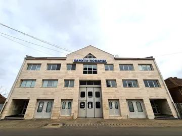 O vedere largă a clădirii SAMGAS ROMANIA, placată cu piatră de culoare deschisă, cu inscripția sub fronton, două rânduri de ferestre deasupra unui rând de uși gri și linii electrice care traversează cerul înnorat.