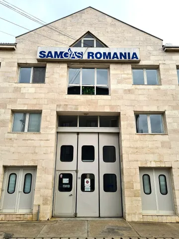 Fațada unei clădiri placate cu piatră, cu inscripția albastră SAMGAS ROMANIA deasupra unei porți mari gri și câte o ușă gri mai mică de fiecare parte.