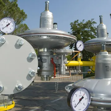 Două regulatoare argintii de presiune pentru gaz, cu carcase rotunde de membrană, manometre și conducte galbene, pe un amplasament în aer liber, cu copaci în spate.
