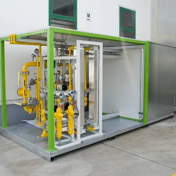 O instalație de gaz cu conducte galbene, robinete și manometre pe o platformă de oțel, sub un cadru verde, amplasată lângă peretele unei clădiri albe.