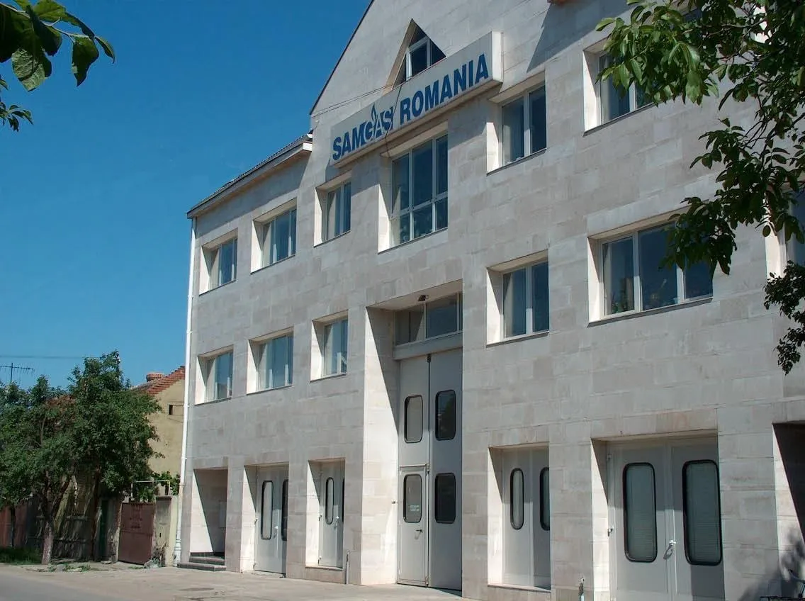 A three-storey stone-clad building with a blue SAMGAS ROMANIA sign on its front, large doors on the ground floor and trees at both sides.