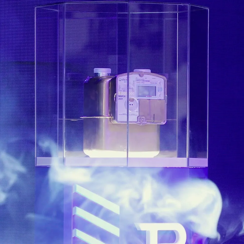A smart gas meter displayed in a glass case on a stage lit in blue, with stage smoke around its base.