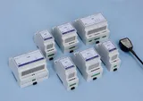 Șapte module albe pentru șină DIN, de dimensiuni diferite, fiecare cu o etichetă albastră și blocuri de borne verzi, așezate pe o suprafață albastru-deschis, lângă un cablu negru terminat cu o mufă mică.