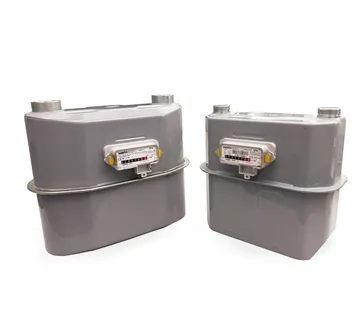 Două contoare de gaz gri de dimensiuni diferite, unul lângă altul, pe fundal alb, fiecare cu o fereastră mică a mecanismului indicator și două racorduri filetate în partea de sus.