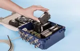 O mână care scoate un modul negru dintr-o carcasă albastră cu capacul deschis, în care se află alte module negre și blocuri de borne verzi, cu conectori de cablu de-a lungul marginii din față.